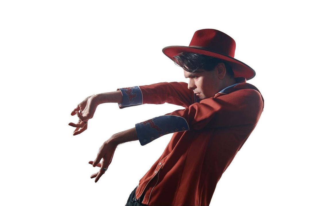 Man in a red cowboy hat and shirt with embroidered cuffs poses on a white background with arms outstretched in a dance-like gesture.
